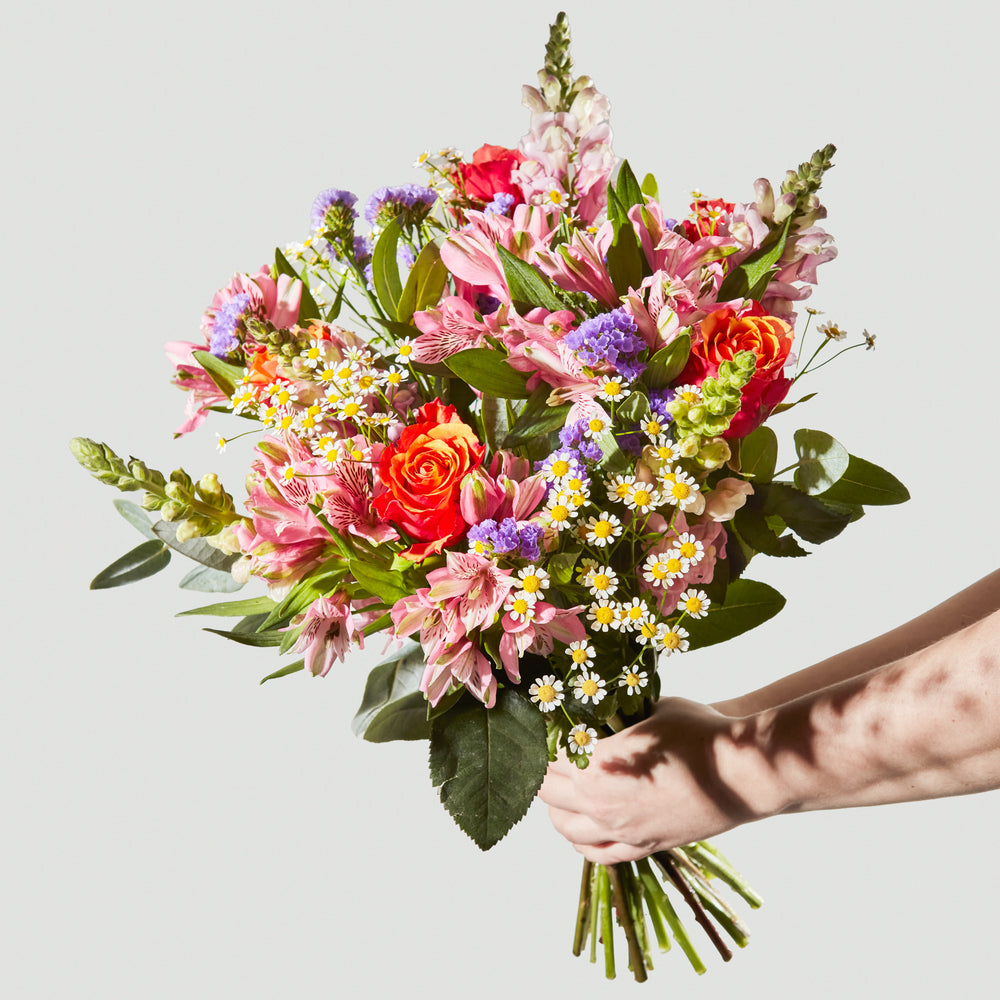 A colorful floral arrangement featuring orange roses, pink alstroemeria, purple statice, and white daisies held by a person.