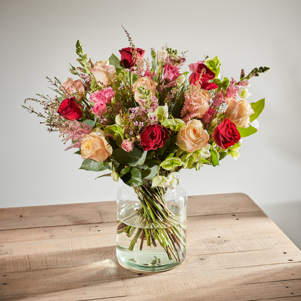 Large floral arrangement with red roses, peach roses, and pink snapdragons in a glass vase.