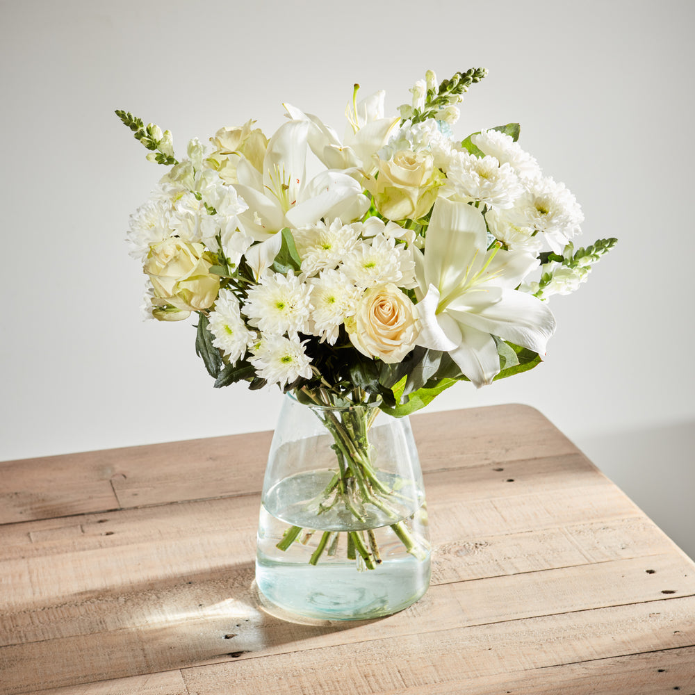 Elegant white floral arrangement with lilies, roses, and chrysanthemums in a clear glass vase.